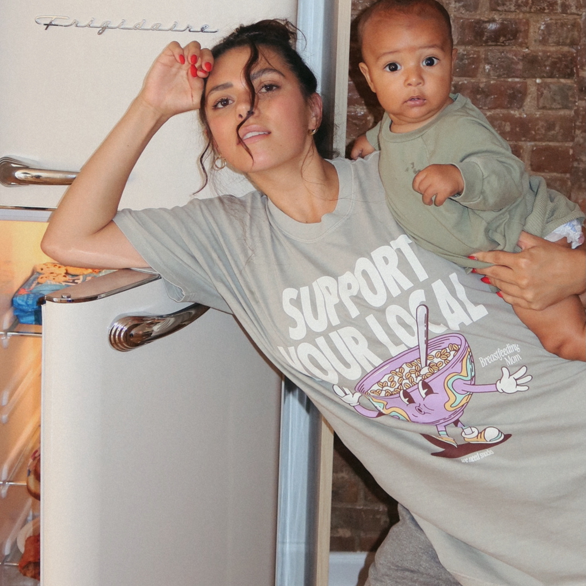 [main] A mom wearing a light sage t-shirt featuring the slogan Support Your Local Breastfeeding Mom and a bright cereal bowl graphic, while holding her son by a fridge.